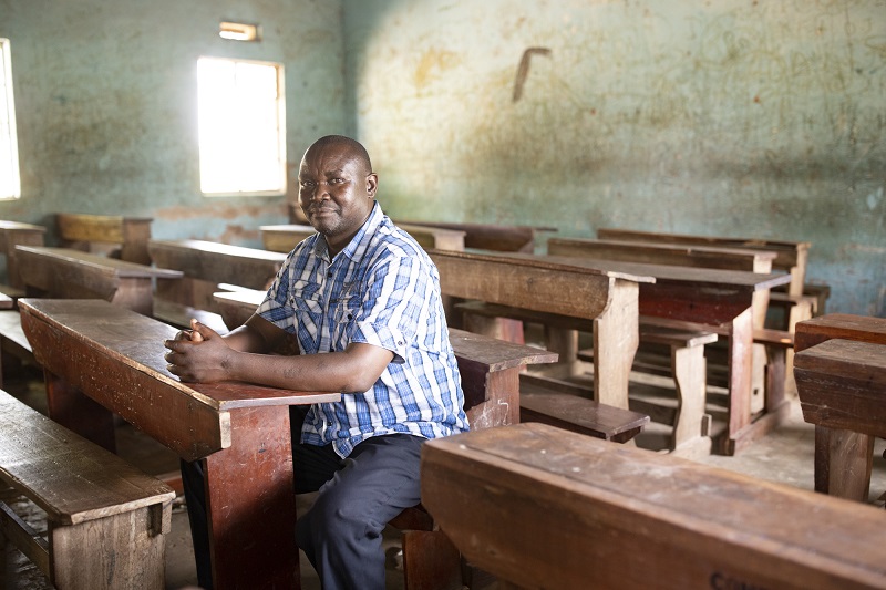A man in Uganda sits in an empty classroom, looking pensively at camera.